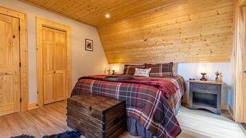Une chambre avec un plafond en bois, un lit recouvert d’une couette à carreaux, une commode et une table de chevet avec une lampe.