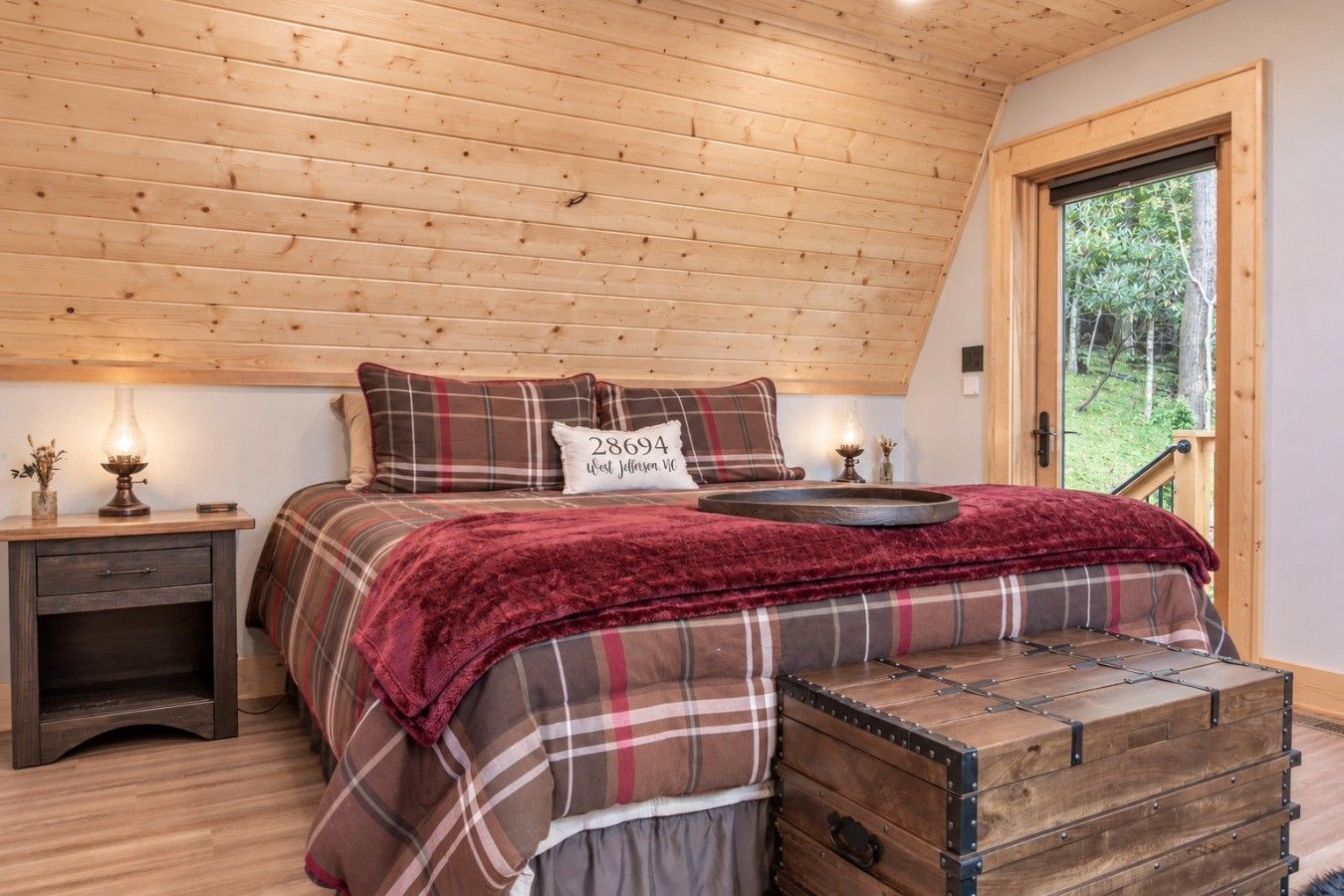 A bedroom with a wooden ceiling, a plaid bedspread, a bedside table with a lamp, and a window with a view of trees.