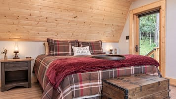 A bedroom with a wooden ceiling, a plaid bedspread, a bedside table with a lamp, and a window with a view of trees.