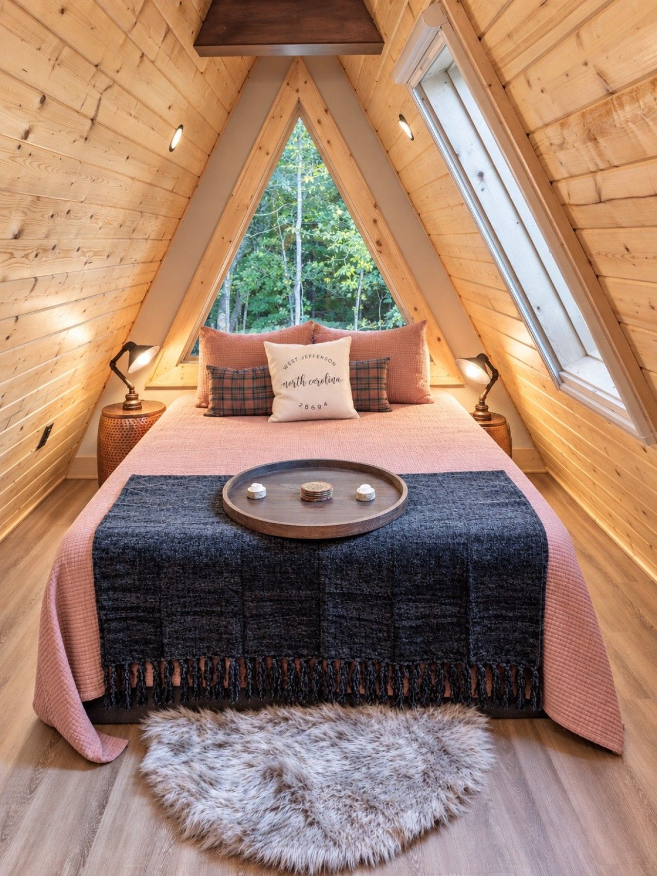 A cozy loft bedroom with a sloped ceiling, a large window, a bed with a decorative pillow, and a small wooden tray on the bed.