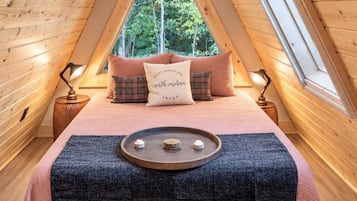 A cozy loft bedroom with a sloped ceiling, a large window, a bed with a decorative pillow, and a small wooden tray on the bed.