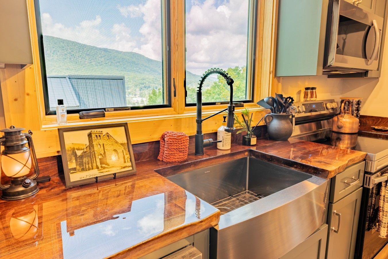 Una cucina con lavello in acciaio inossidabile, piano di lavoro in legno, un quadro incorniciato, un bollitore e una finestra con vista su un edificio e delle colline.