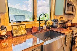 Una cucina con lavello in acciaio inossidabile, piano di lavoro in legno, un quadro incorniciato, un bollitore e una finestra con vista su un edificio e delle colline.