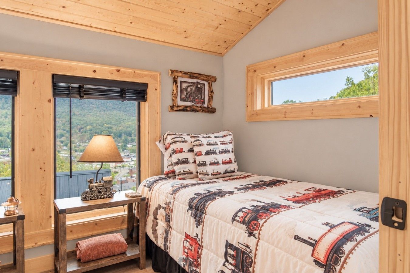 A bedroom with a bed, a bedside table with a lamp, a window with a view, and a wooden ceiling.