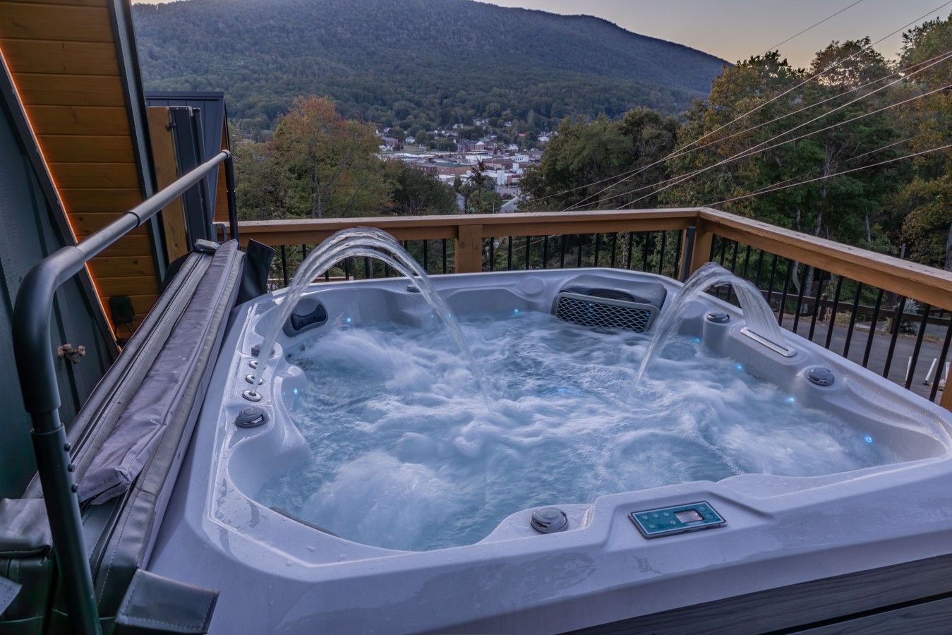 A hot tub with a wooden deck and railing, surrounded by a mountainous landscape.
