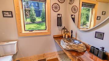 A bathroom with a stone basin, wooden floor, and a window with a view of a house and trees.