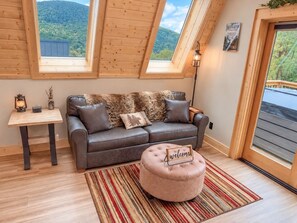 Ein gemütliches Zimmer mit einer Couch, einem Hocker und einem kleinen Tisch mit Lampe. Über der Couch befinden sich Dachfenster und eine Tür führt nach draußen.