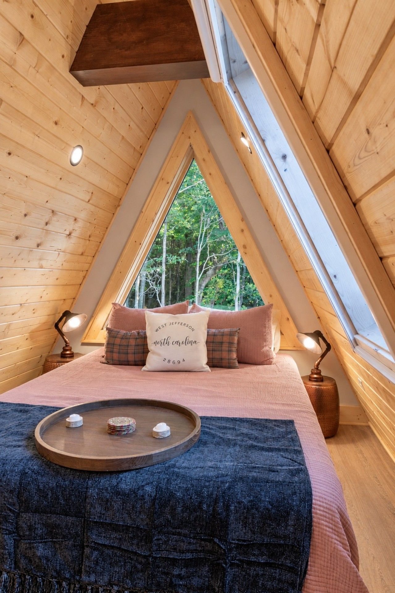 A cozy attic bedroom with a sloped ceiling, a large window, a bed with a decorative pillow, and a bedside table with a lamp.