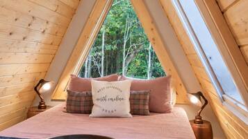 A cozy attic bedroom with a sloped ceiling, a large window, a bed with a decorative pillow, and a bedside table with a lamp.