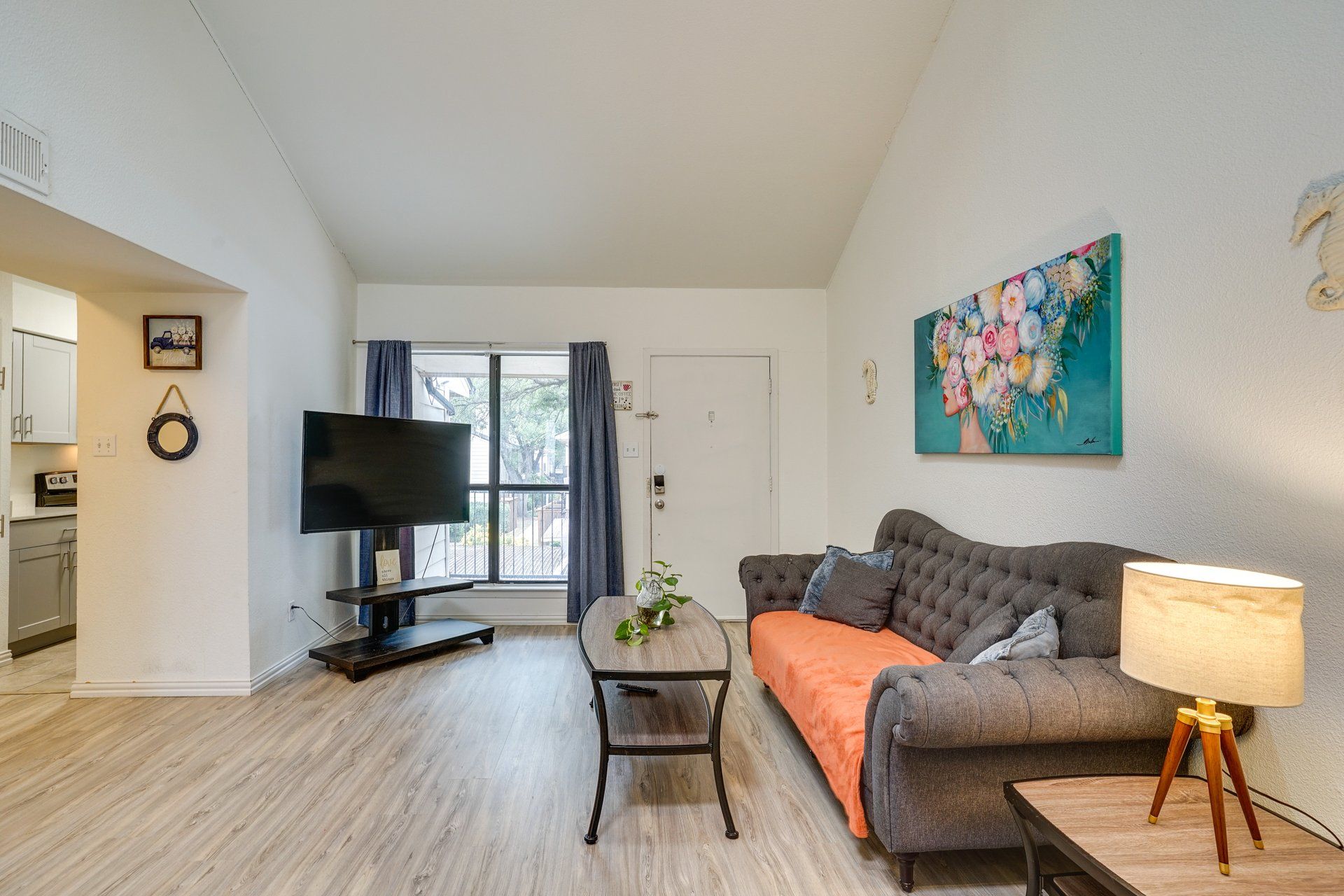 A living room with a flat-screen TV, a grey sofa, a small table with a plant, and a painting on the wall.