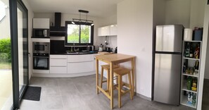 A modern kitchen with a built-in oven, microwave, and refrigerator, a wooden table, and a window with a view of greenery. - A Stay in Le Loc'h, Plogoff, Finistère, Brittany (Plogoff)