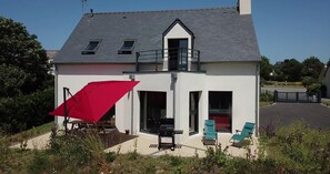 A two-story house with a balcony, a red umbrella, and two green lounge chairs.