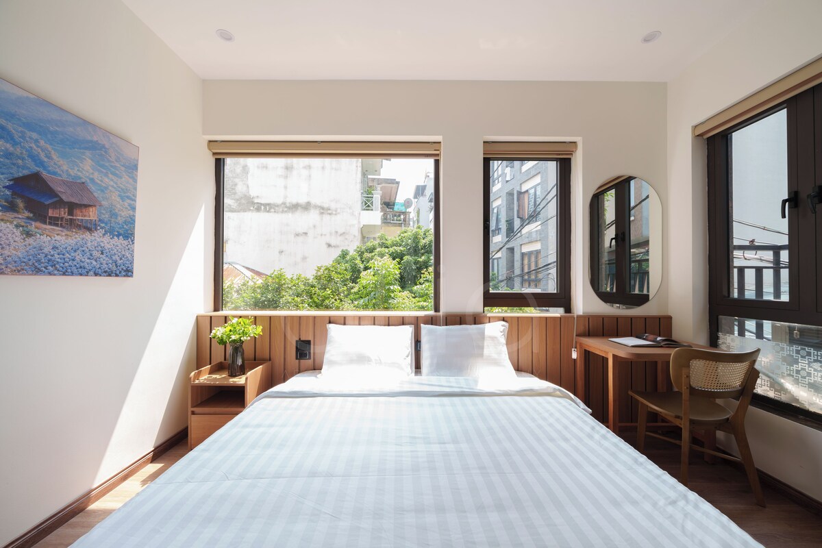 Un dormitorio moderno con una cama grande, una mesita de noche de madera, una pequeña mesa, una silla y un amplio ventanal con vistas a edificios y árboles.