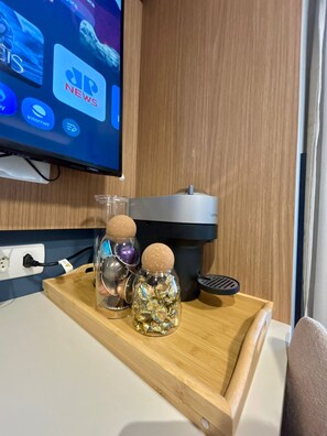 A wooden tray with two glass jars containing wrapped candies and a metal container, placed on a table with a power outlet and a television displaying a news channel. - Level Brooklin 1217 (São Paulo)