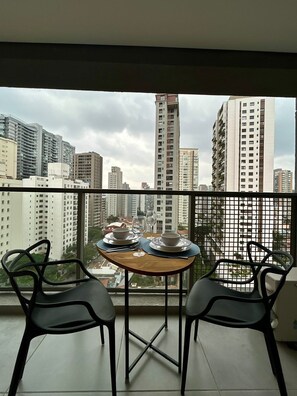 A balcony with two chairs, a table set with plates, bowls, and glasses, overlooking a cityscape with high-rise buildings. - Level Brooklin 1217 (São Paulo)