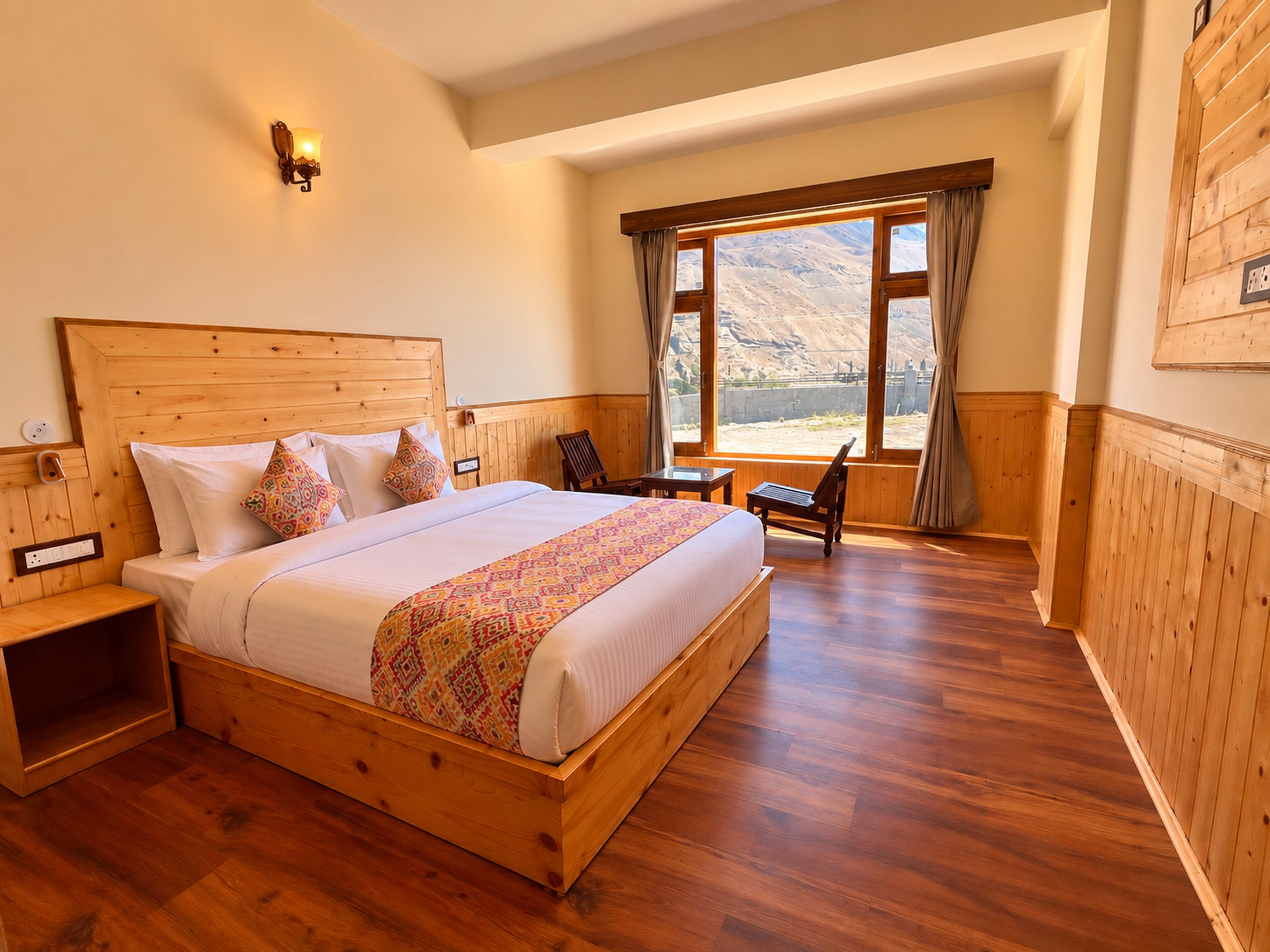 Una cama con estructura de madera y colcha estampada, una mesita de noche, una silla y una ventana con vistas a un paisaje montañoso.