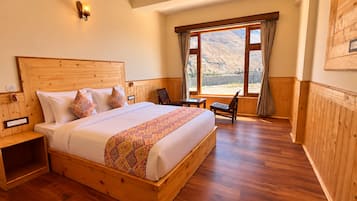 Una cama con estructura de madera y colcha estampada, una mesita de noche, una silla y una ventana con vistas a un paisaje montañoso.