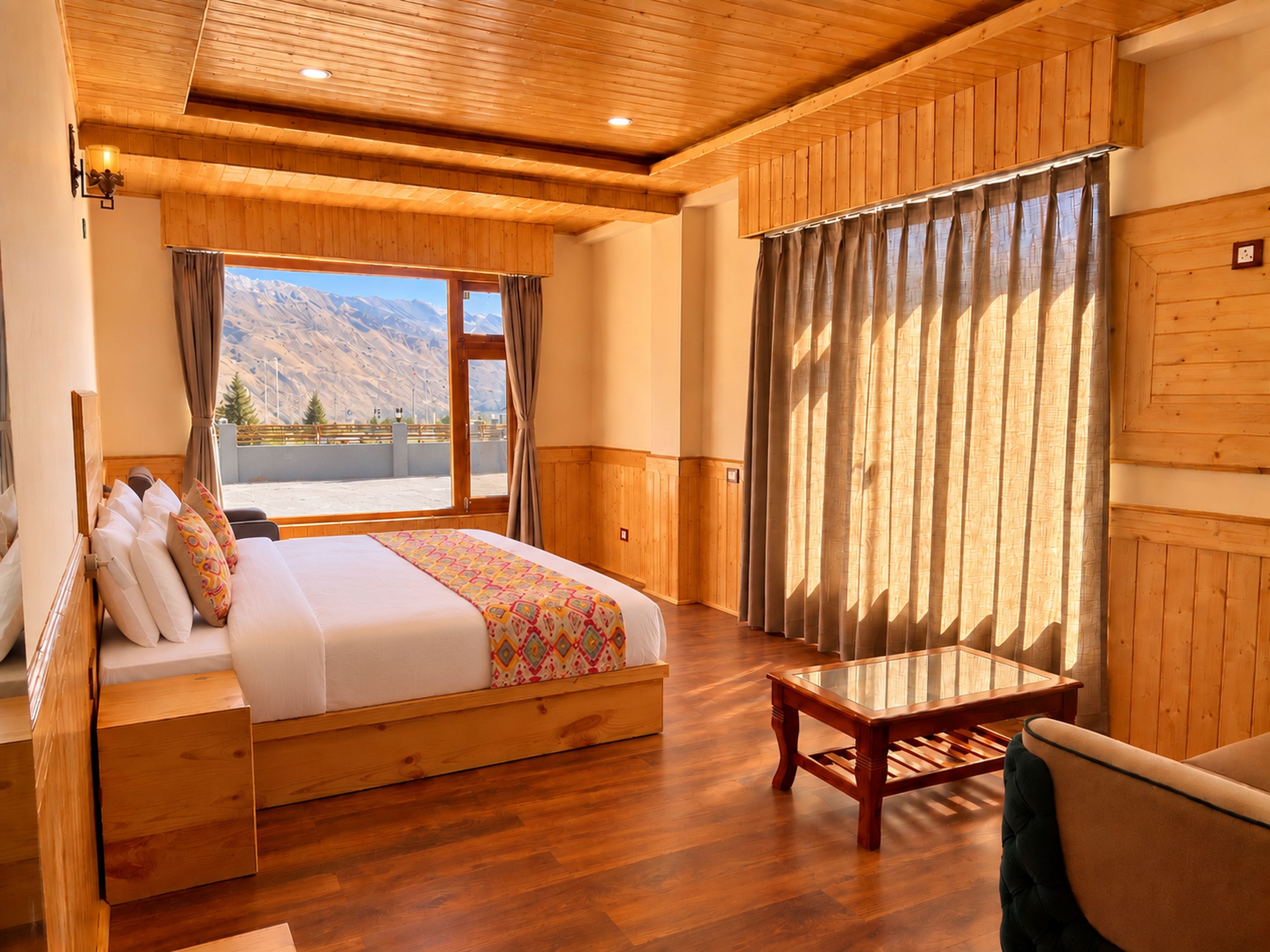 Una habitación con techo de madera, cama con manta estampada, mesa de madera y vista a montañas a través de una ventana.