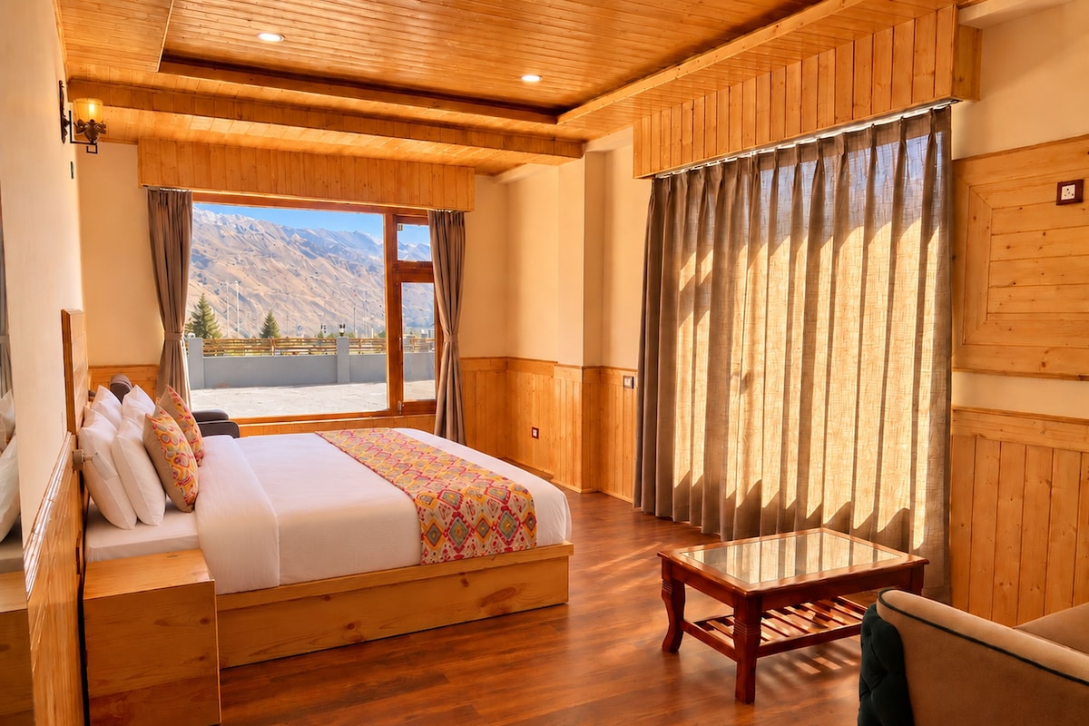 Una stanza con soffitto in legno, un letto con coperta a motivi decorativi, un tavolo di legno e vista sulle montagne attraverso una finestra.
