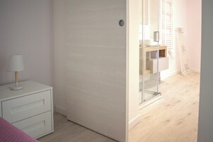 Una habitación con un aparador blanco, un vidrio que da al baño y piso de madera.