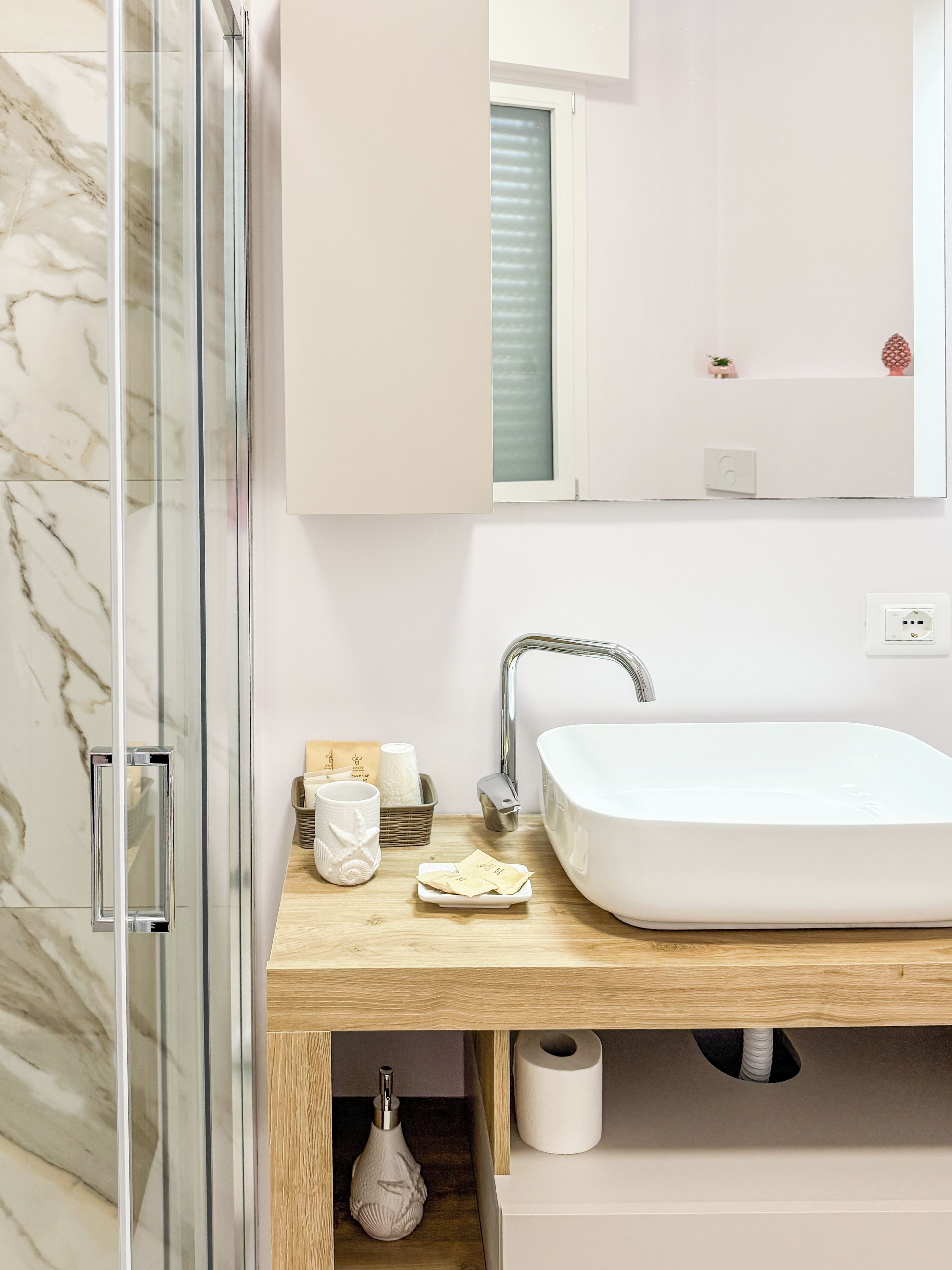 A bathroom with a glass shower enclosure, a white sink with a modern faucet, and a wooden vanity with toiletries.