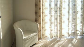Un rincón de una habitación con un sillón blanco, suelo de madera y una ventana con cortinas traslúcidas.