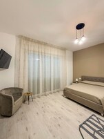 Un dormitorio con cama, una silla, una mesita y un televisor colocado en la pared.
