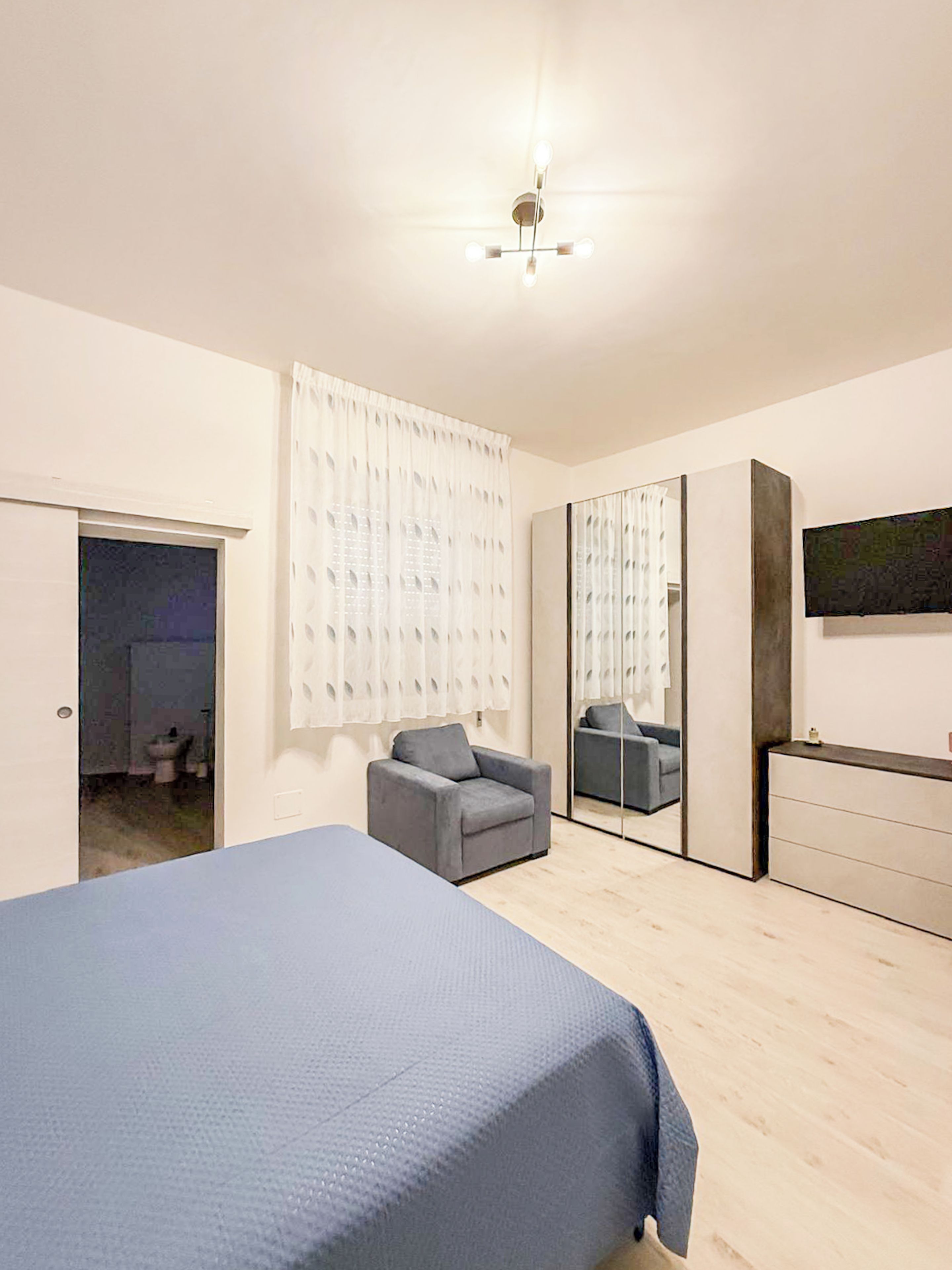 A bedroom with a bed, a chair, a dresser, a mirror, and a ceiling light fixture.
