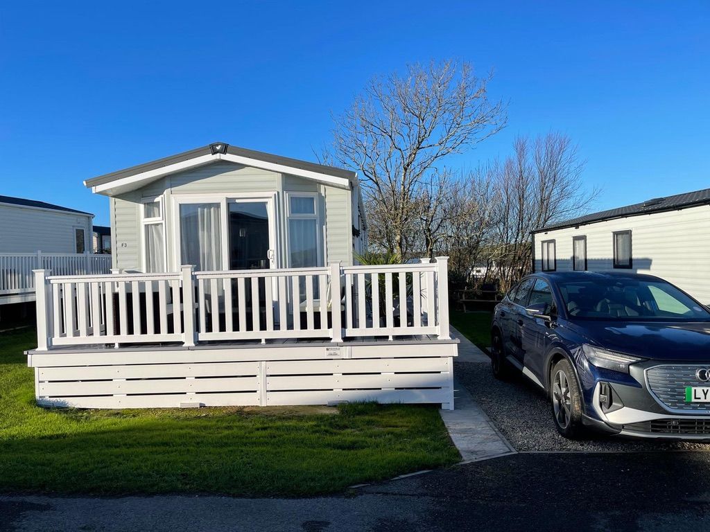 Une maison mobile à l’extérieur blanc, dotée d’une terrasse et avec une voiture garée devant.