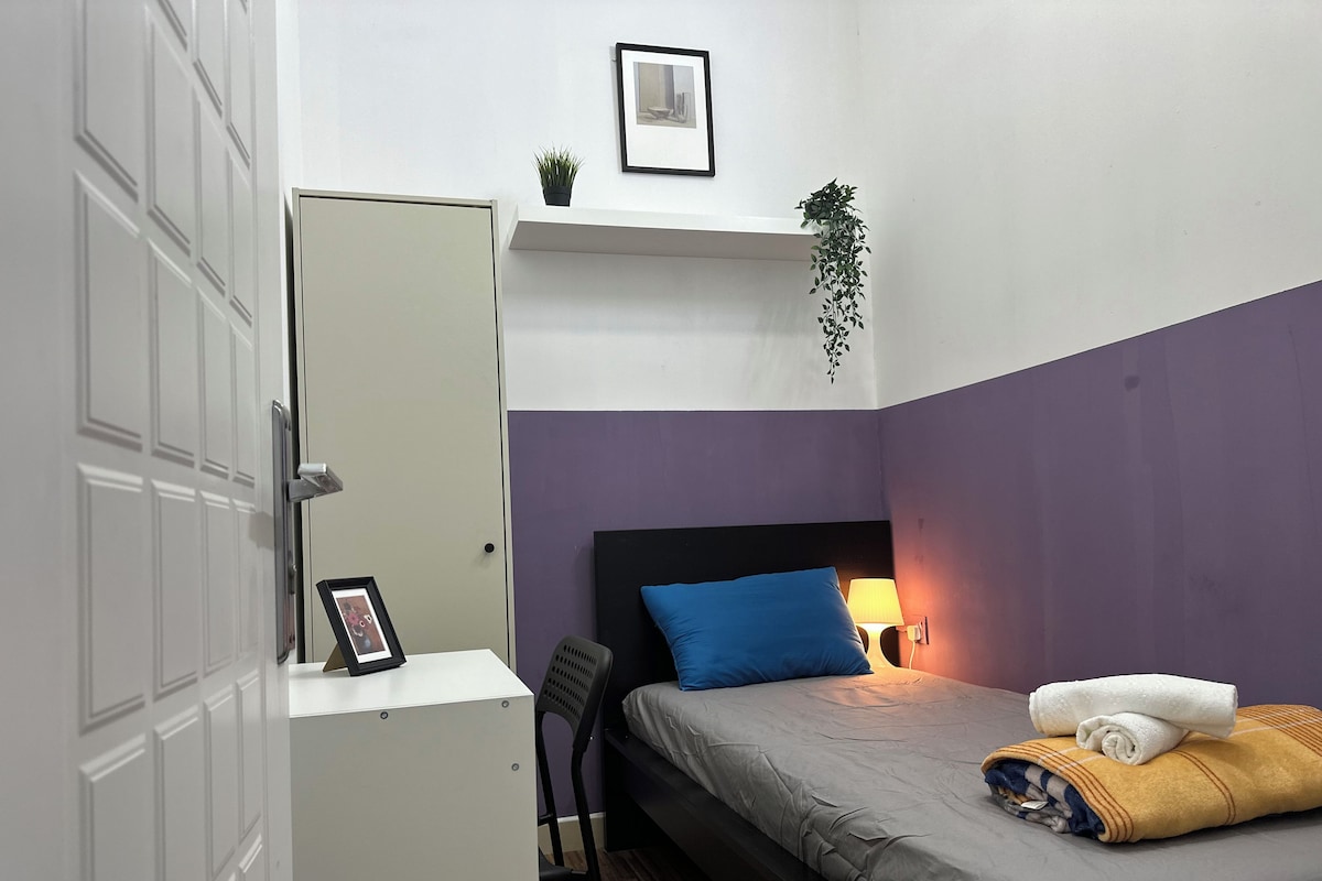 A compact room with a bed, a desk, a chair, a bedside lamp, and a door.