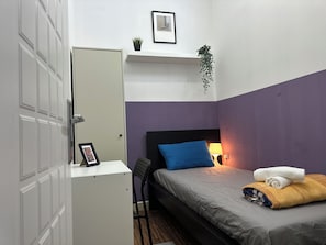 A compact room with a bed, a desk, a chair, a bedside lamp, and a door.