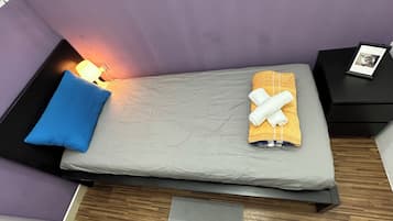 Una cama individual con un colchón gris, una almohada azul y una toalla doblada sobre otra naranja. Hay una mesita de noche con una lámpara y una foto enmarcada. La habitación tiene paredes moradas y piso de madera.