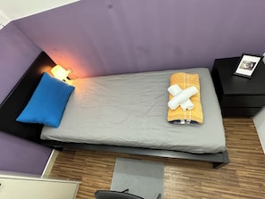 A single bed with a grey mattress, a blue pillow, and a folded towel on an orange towel. There is a nightstand with a lamp and a framed photo. The room has purple walls and a wooden floor. - Fancy Partition Room in Al Barsha1 (Dubai)