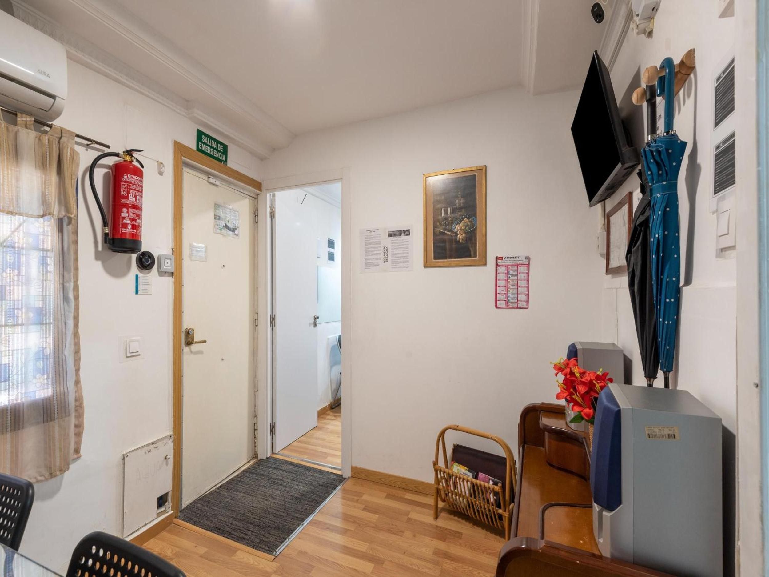 Ett hotellrum med en brandsläckare, en dörr med en nödutgångsskylt, en inramad tavla på väggen, en tv och ett blått paraply.
