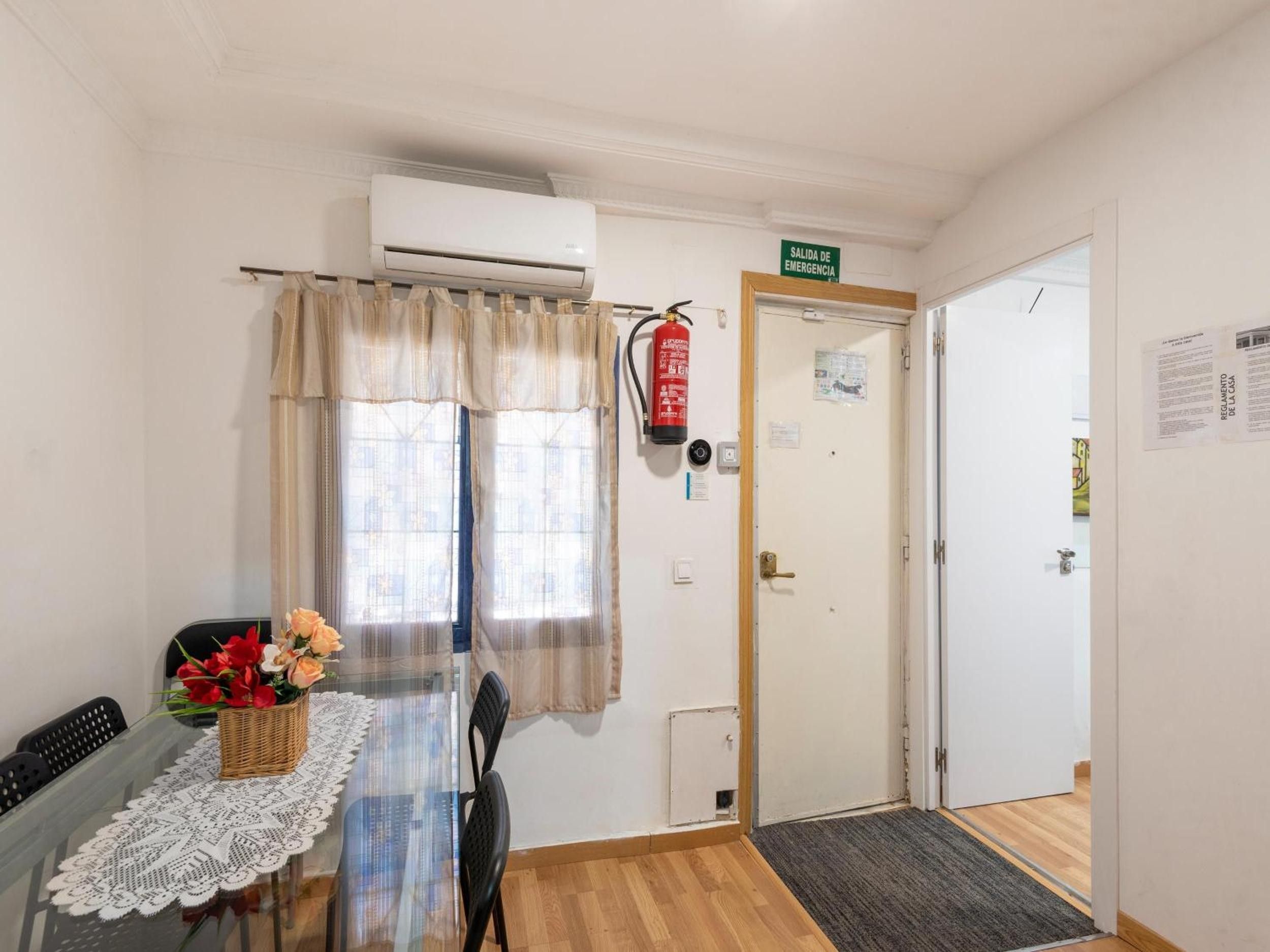 Una habitación con una mesa de comedor, sillas, un extintor, una puerta con letrero de salida de emergencia y una ventana con cortinas translúcidas.