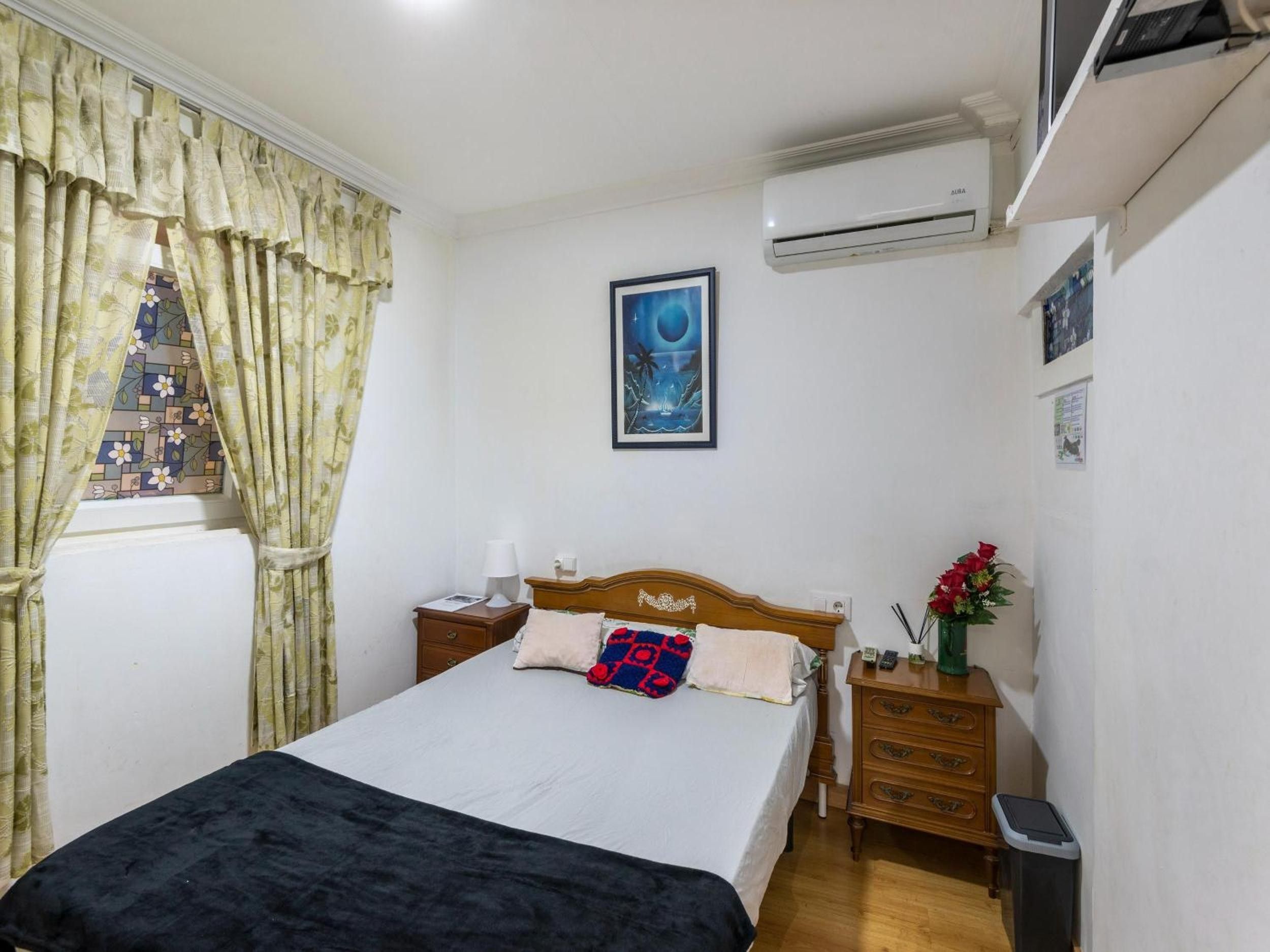 Une chambre à coucher comprenant un lit, une table de chevet, une commode, un appareil de climatisation et une fenêtre avec des rideaux.