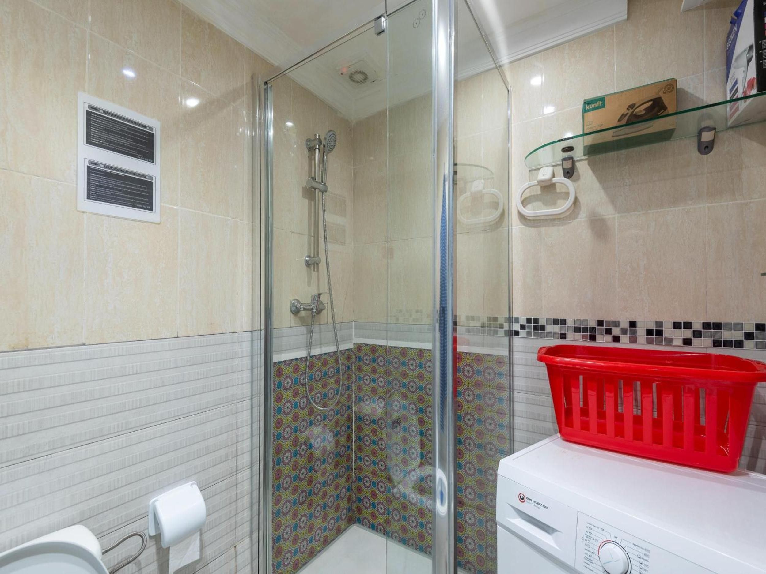 Un bagno con cabina doccia in vetro, un water, una lavatrice e un cesto della biancheria rosso.