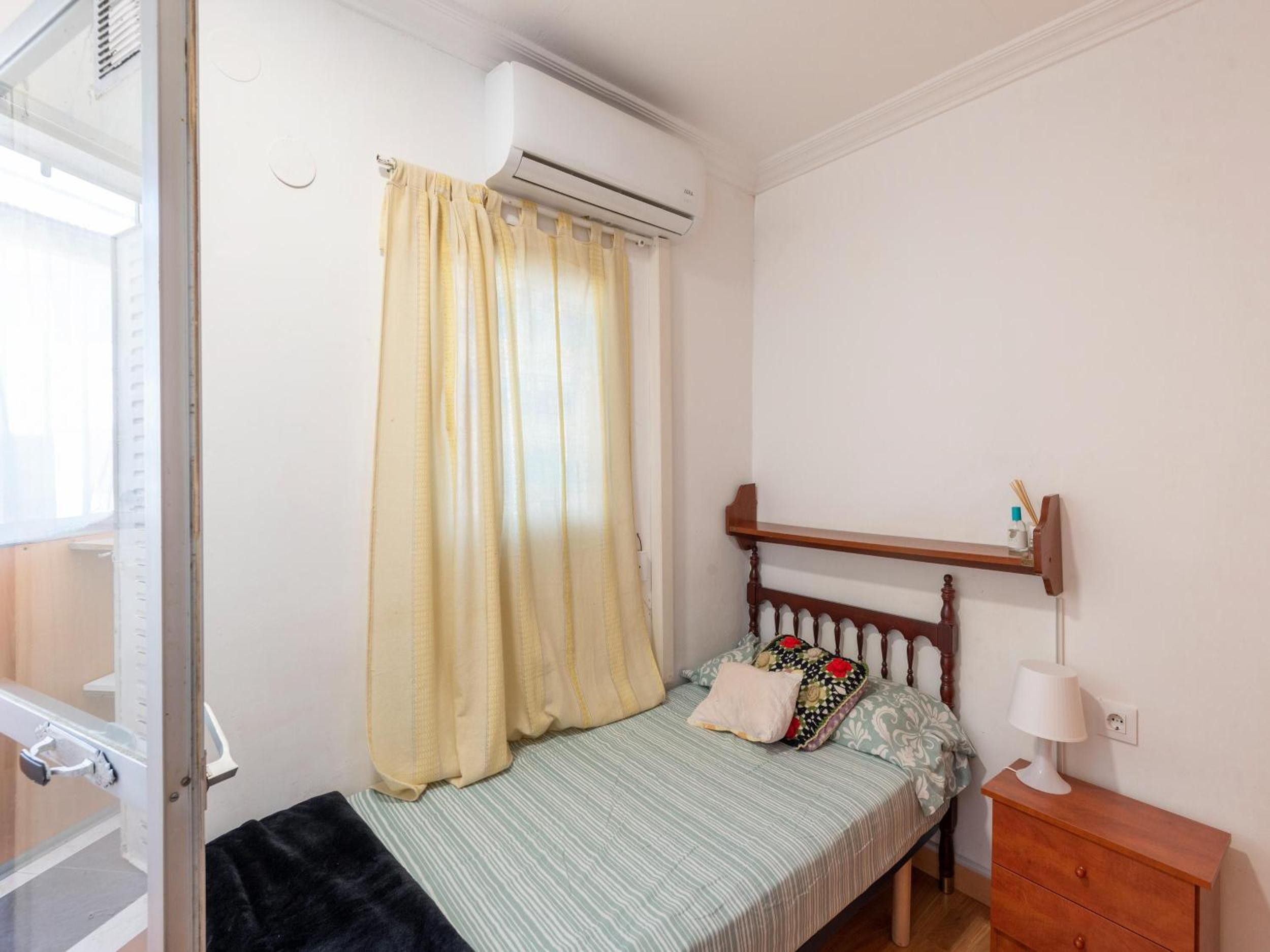 A bedroom with a bed, a nightstand, a lamp, a shelf with items, and an air conditioning unit.