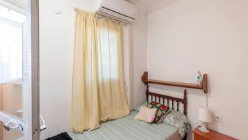 A bedroom with a bed, a nightstand, a lamp, a shelf with items, and an air conditioning unit.