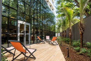 An outdoor seating area with modern chairs and a wooden deck, surrounded by glass walls and tropical plants. - On Paulista 817 (Vila Mariana)