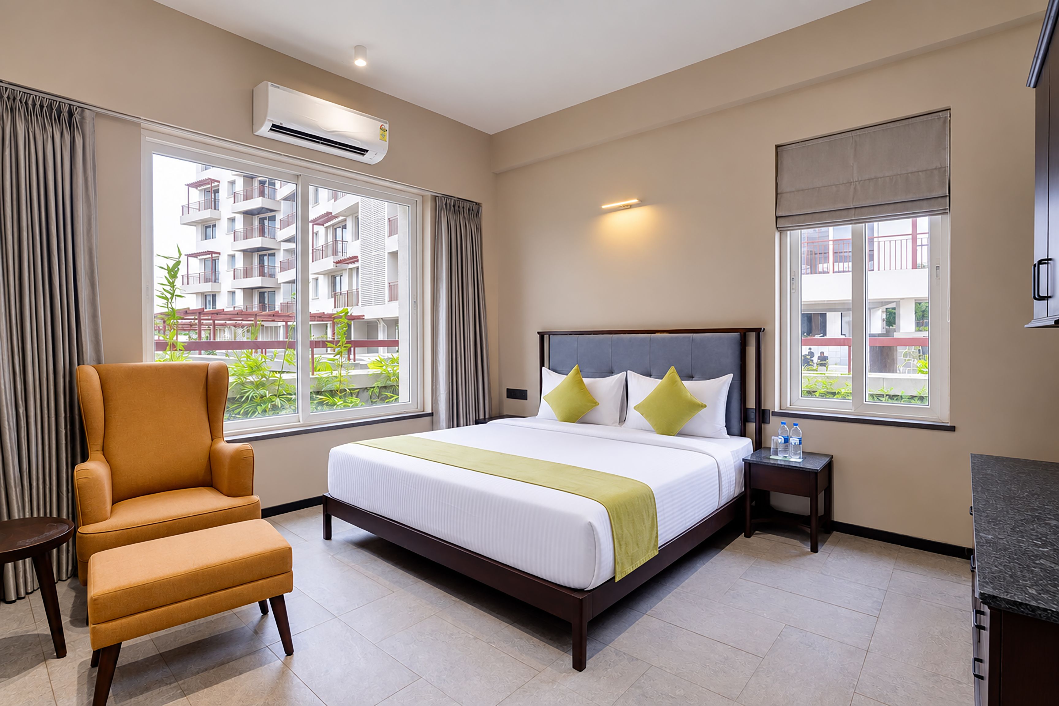 Quarto de hotel com cama, cadeira, mesa de cabeceira e janela com vista para um edifício.