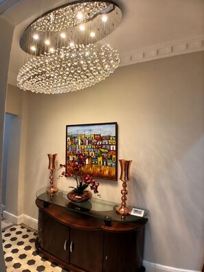Ein Zimmer mit einem dekorativen Chandelier, einem Konsolentisch mit Glasplatte und einer Blumenvase sowie einem farbenfrohen Gemälde an der Wand.