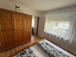 Habitació amb un armari de fusta, un llit amb roba de llit estampada, una finestra amb cortines transparents i vista cap a l'exterior.