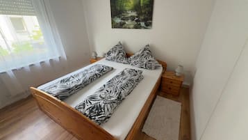 Ein Schlafzimmer mit einem hölzernen Bett, einem Waldgemälde, einem Fenster mit Jalousien und einem Nachttisch mit Lampe.