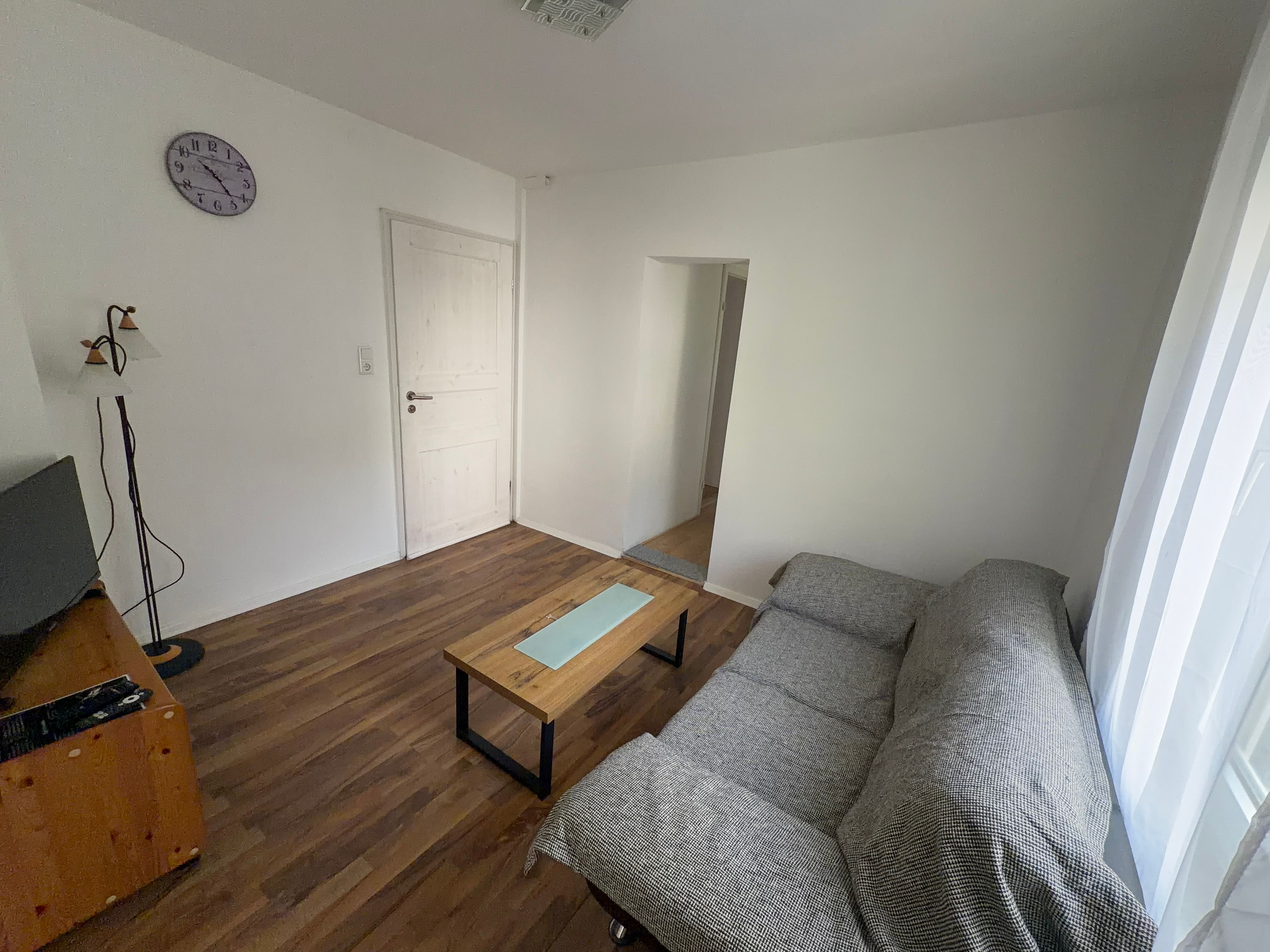 Ein Wohnzimmer mit Holzboden, einer Couch, einem kleinen Tisch, einer Wanduhr und einer Tür zu einem anderen Raum.