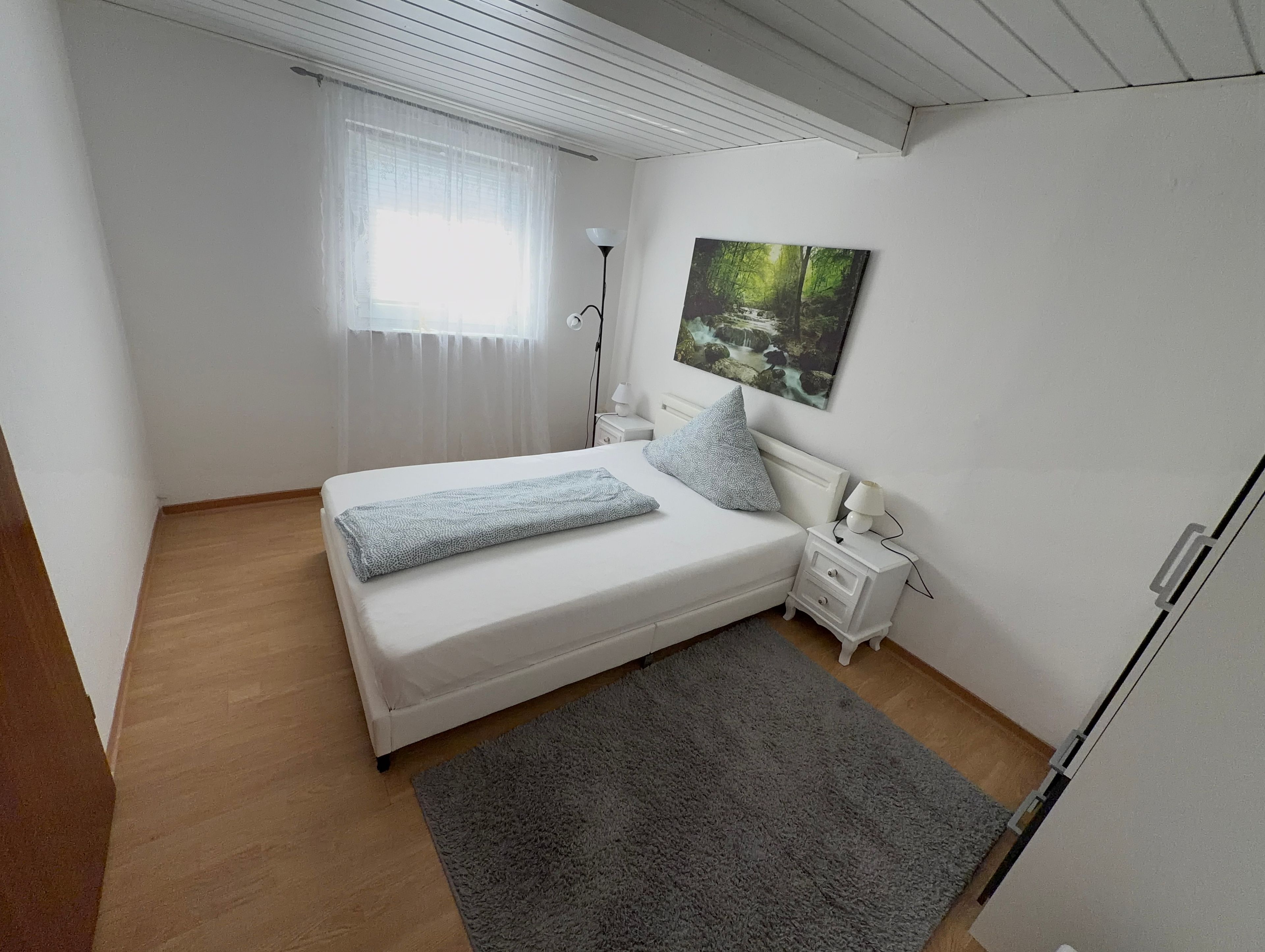 Un dormitorio con una cama, mesitas de noche, una lámpara, un cuadro y una ventana con cortinas.