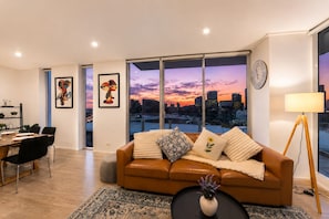Ein modernes Wohnzimmer mit Blick auf die Stadtlandschaft, ein braunes Ledersofa, ein Essbereich mit Tisch und Stühlen und ein großes Fenster.