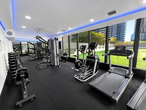 Un gimnasio con diversos equipos de ejercicio, ventanas amplias y vistas a un área verde exterior.