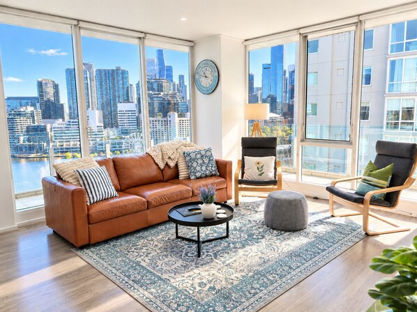 A spacious living room with a large window offering a city view, a leather sofa, a coffee table, and two chairs. - Spectacular City Views | Large 3BR 2BA Near Crown & MCEC (Southbank)