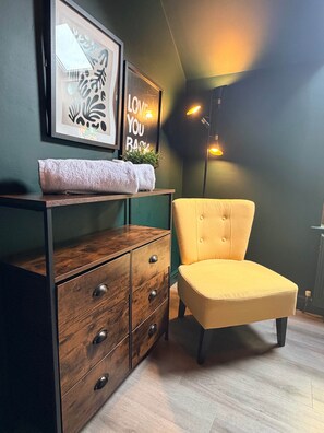 Ein Zimmer mit einem hölzernen Schminktisch, einem gelben Stuhl und Wandbildern, darunter ein gerahmtes Bild und ein Schild.